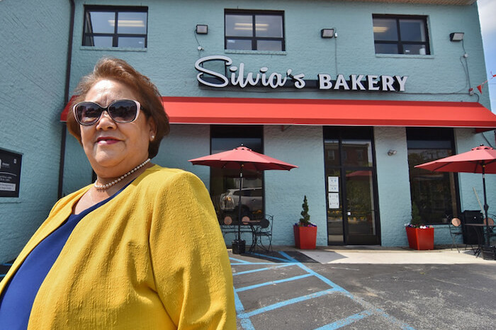 Silvia, owner of Silvia's Bakery, standing in front of her bakery with a red awning and tables with red umbrellas